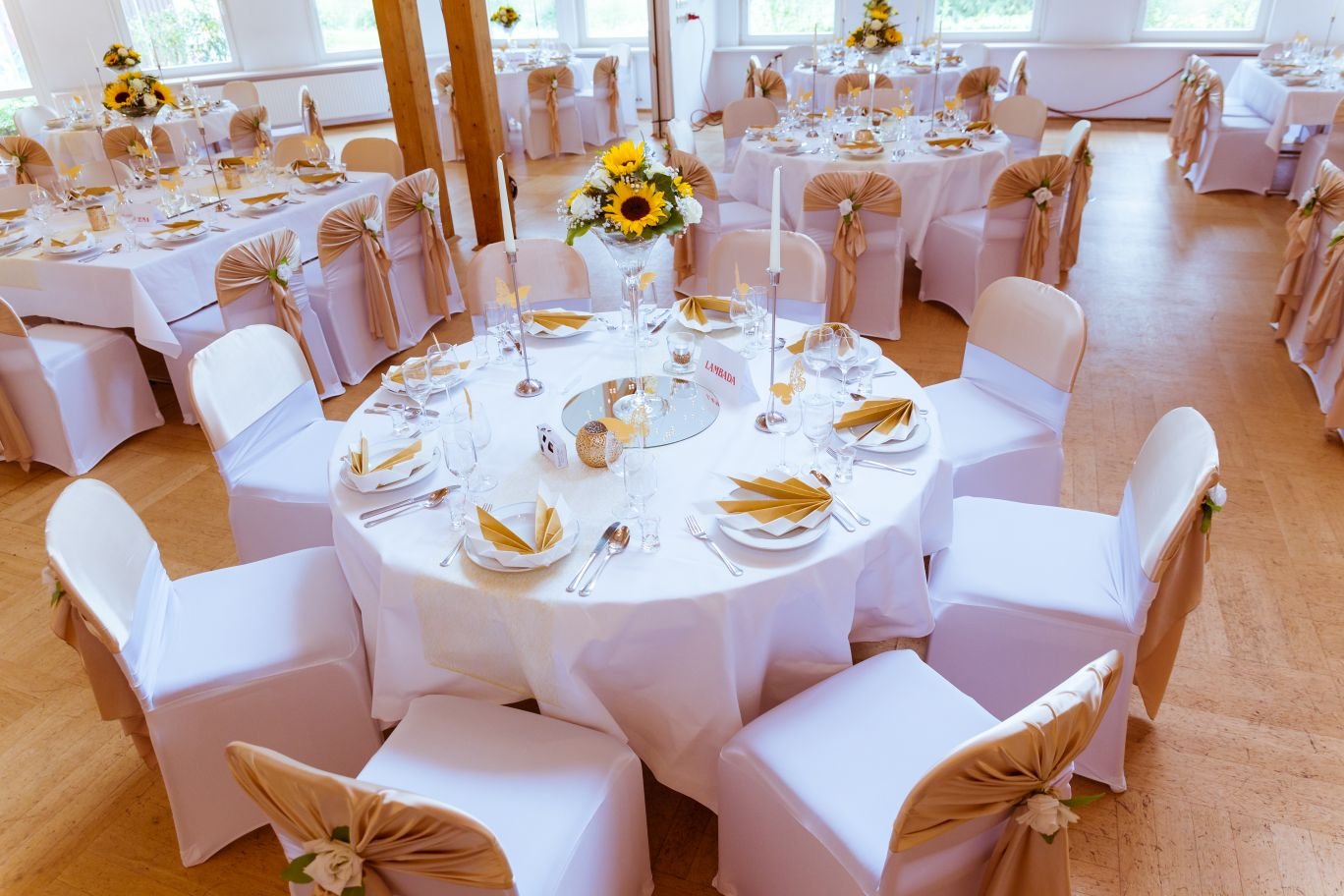 Festsaal mit runden Tischen, weißen Stuhlhussen und goldenen Schleifen mit Sonnenblumen